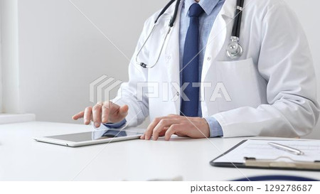 Doctor man wearing white medical coat and stethoscope using digital tablet while sitting at desk in medical office, with medical chart and pen in the foreground. Medicine concept 129278687