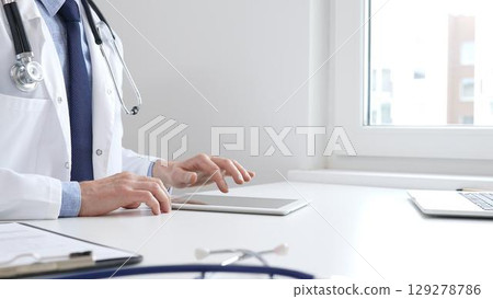 Male doctor wearing lab coat and stethoscope using digital tablet while sitting at desk in medical office, with medical chart and pen in the foreground Male doctor wearing lab coat and stethoscope using digital tablet while sitting at desk in medical office, with medical chart and pen in the foreground 129278786