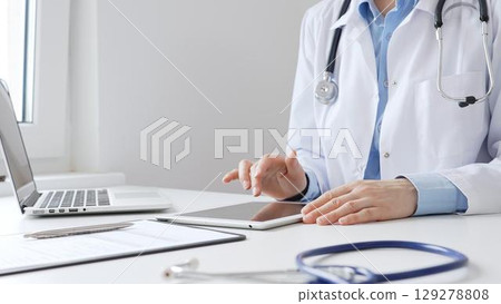 Female doctor wearing white medical coat and stethoscope using digital tablet with laptop and medical records on desk in medical office, showcasing modern healthcare technology. Medicine and helthcare 129278808