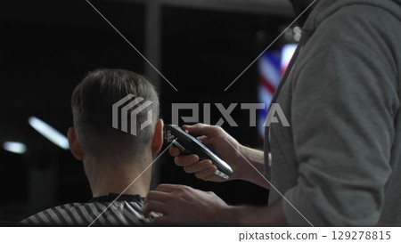 Skilled barber trimming client's hair with electric razor, creating precise contemporary hairstyle inside sleek urban barbershop setting 129278815