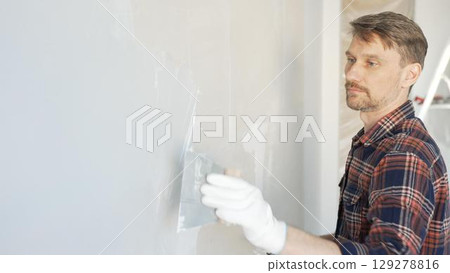 Male construction worker spreading plaster on white wall, wearing protective gloves during home renovation project with putty knife Male construction worker spreading plaster on white wall, wearing protective gloves during home renovation project with putty knife 129278816