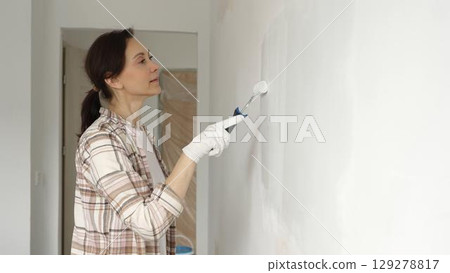Middle aged woman wearing protective gloves painting white wall with paint roller, updating interior and giving room clean, modern look during home renovation project 129278817