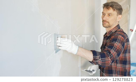 Male construction worker spreading plaster on white wall, wearing protective gloves during home renovation project with putty knife 129278823