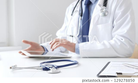 Male doctor wearing white medical coat and stethoscope is using digital tablet while sitting at desk in medical office, with medical chart and pen in the foreground. Medicine and health care concept Male doctor wearing white medical coat and stethoscope is using digital tablet while sitting at desk in medical office, with medical chart and pen in the foreground. Medicine and health care concept 129278850