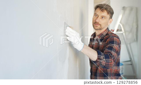 Construction worker man wearing gloves and holding a putty knife is applying plaster on a white wall, performing home renovation and improvement Construction worker man wearing gloves and holding a putty knife is applying plaster on a white wall, performing home renovation and improvement 129278856