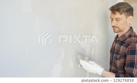 Construction worker man wearing gloves and holding a putty knife is applying plaster on a white wall, performing home renovation and improvement 129278858