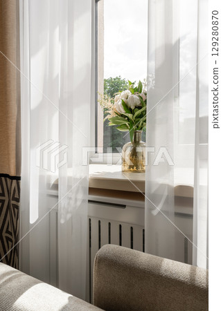 Sunlit Window with Sheer Curtains and Vase of White Flowers 129280870