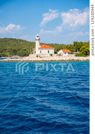 Stone lighthouse with red roof on the rocky coast of Hvar Island, Croatia, surrounded by pine trees and clear blue sea. Navigation, solitude and maritime heritage in the Adriatic. 129280944