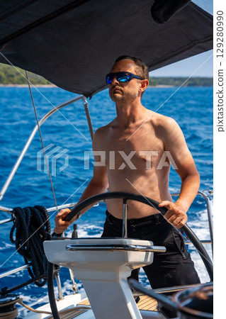 Man with bare torso steering a yacht on the open sea on a sunny day. Concept: summer vacation, yachting, sea travel. Man with bare torso steering a yacht on the open sea on a sunny day. Concept: summer vacation, yachting, sea travel. 129280990
