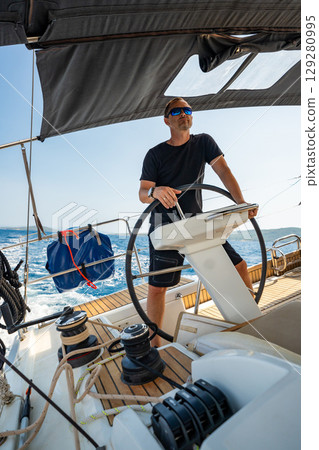 Man standing at the helm of a sailing yacht near Croatia, steering the boat through open water. Leadership, control, sailing lifestyle and freedom at sea. Man standing at the helm of a sailing yacht near Croatia, steering the boat through open water. Leadership, control, sailing lifestyle and freedom at sea. 129280995