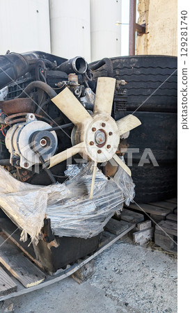 Engine cooling fan assembly with attached components, resting on wooden pallets beside large tires, showcasing industrial machinery and mechanical parts in a storage environment Engine cooling fan assembly with attached components, resting on wooden pallets beside large tires, showcasing industrial machinery and mechanical parts in a storage environment 129281740