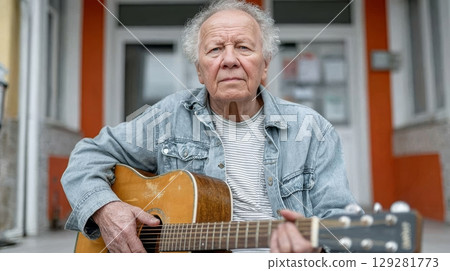 Elderly man playing acoustic guitar outdoors, wearing denim jacket, showcasing musical talent and passion in a vibrant urban setting 129281773