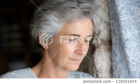 Mature woman with gray hair is thoughtfully gazing out of a window, reflecting on life experiences, showcasing a moment of contemplation and serenity 129281854