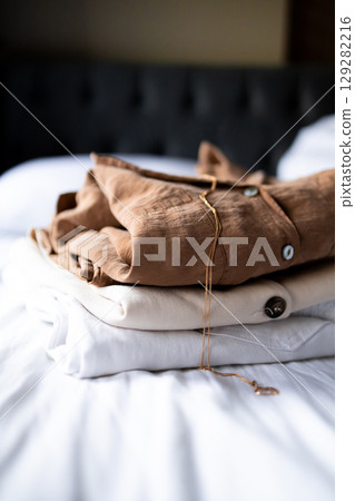Neatly folded brown, beige, and white clothes on bed with necklace Neatly folded brown, beige, and white clothes on bed with necklace 129282216