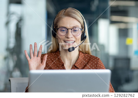 A woman with a headset, wearing glasses, waves while looking at a laptop screen in a modern office setting. A woman with a headset, wearing glasses, waves while looking at a laptop screen in a modern office setting. 129282762