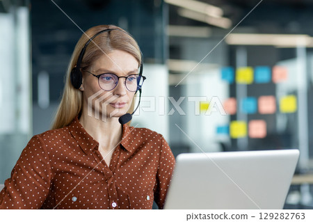 A female customer service representative wears a headset while working on her laptop in a modern office setting. A female customer service representative wears a headset while working on her laptop in a modern office setting. 129282763