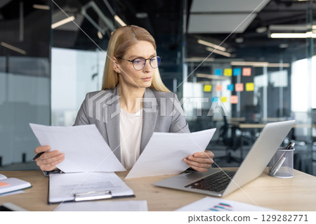 A focused businesswoman in a modern office setting reviewing documents, with a laptop nearby, illustrating work and professionalism. 129282771