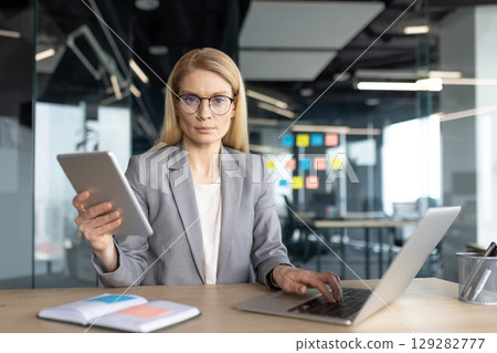 A woman in a business suit uses a laptop and tablet in a modern office setting, possibly working in management or an executive role. 129282777