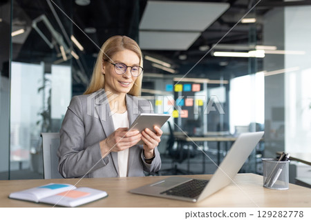 A professional woman in a modern office setting, focused on her tablet, with a laptop and office supplies nearby. 129282778