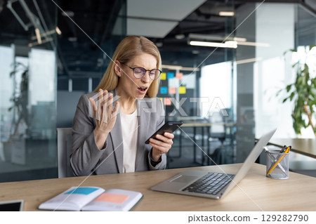 A businesswoman is experiencing a stressful situation while looking at her phone in the office. 129282790