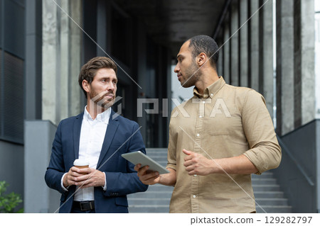 Two business professionals discuss work while looking at a tablet outside a modern building. One holds a coffee cup. 129282797