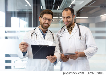 Two doctors, in white coats with stethoscopes, review paperwork and a tablet in a modern office setting. They appear focused and collaborative. 129282806