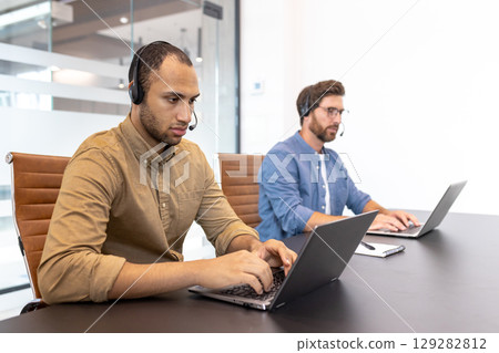 Two diverse call center agents work on laptops, wearing headsets in a modern office environment, providing customer support. Two diverse call center agents work on laptops, wearing headsets in a modern office environment, providing customer support. 129282812