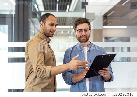 Two diverse business professionals discuss documents in a modern office setting, possibly during a collaborative meeting or presentation. Two diverse business professionals discuss documents in a modern office setting, possibly during a collaborative meeting or presentation. 129282813