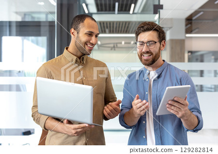 Two smiling men collaborate, one with a laptop, the other with a tablet, in a modern office setting. 129282814