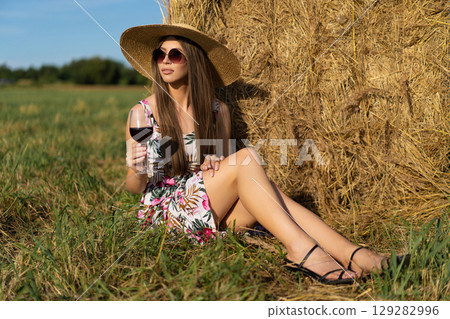 beautiful glamorous girl in a dress and a straw hat on a field near a haystack with a glass of red wine. beautiful glamorous girl in a dress and a straw hat on a field near a haystack with a glass of red wine. 129282996