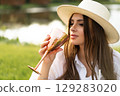 beautiful glamorous girl in a white dress and a straw hat on the shore of the lake with a goblet in her hands. 129283020