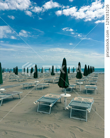 Closed umbrellas and sun loungers on a sandy beach against the backdrop of the sea coast. Empty beach. Closed umbrellas and sun loungers on a sandy beach against the backdrop of the sea coast. Empty beach. 129283216