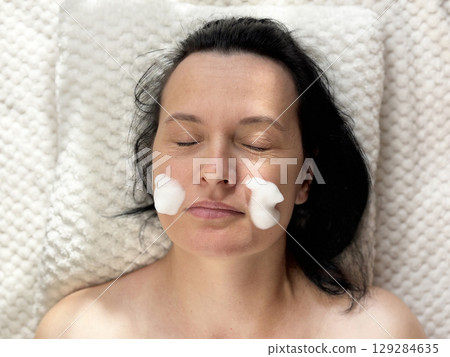 Mature woman lying on a cosmetology table during professional foam facial cleansing procedure. Anti aging skincare treatment in beauty clinic. Wrinkles, skin care, beauty and wellness concept 129284635