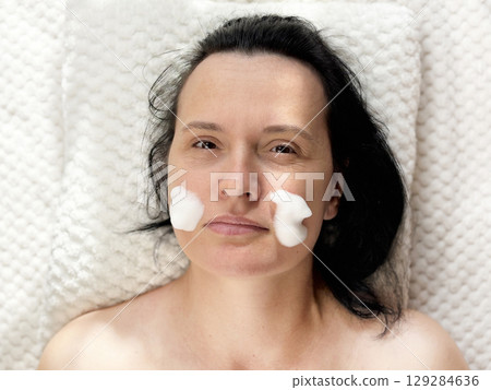 Mature woman lying on a cosmetology table during professional foam facial cleansing procedure. Anti aging skincare treatment in beauty clinic. Wrinkles, skin care, beauty and wellness concept Mature woman lying on a cosmetology table during professional foam facial cleansing procedure. Anti aging skincare treatment in beauty clinic. Wrinkles, skin care, beauty and wellness concept 129284636