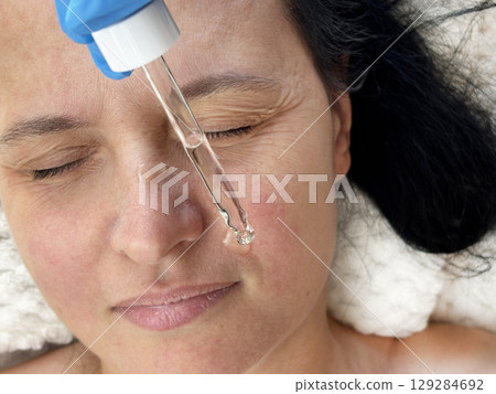 Mature woman with wrinkles undergoing cosmetic skincare procedure. Cosmetologist applying transparent moisturizing serum on her face. Anti aging treatment, beauty and self care for mature skin Mature woman with wrinkles undergoing cosmetic skincare procedure. Cosmetologist applying transparent moisturizing serum on her face. Anti aging treatment, beauty and self care for mature skin 129284692