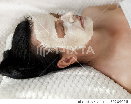 Mature woman with wrinkles lying on a white cosmetology table during a facial procedure. White clay cosmetic mask is applied on her face. Skin care for aging skin. Beauty and spa treatment Mature woman with wrinkles lying on a white cosmetology table during a facial procedure. White clay cosmetic mask is applied on her face. Skin care for aging skin. Beauty and spa treatment 129284696