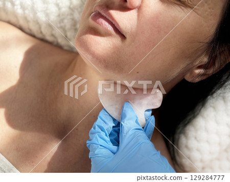 Mature woman lying on cosmetology table during professional facial massage with natural scraper. Anti aging skin care in beauty clinic. Wrinkles, skin care, beauty and wellness concept 129284777