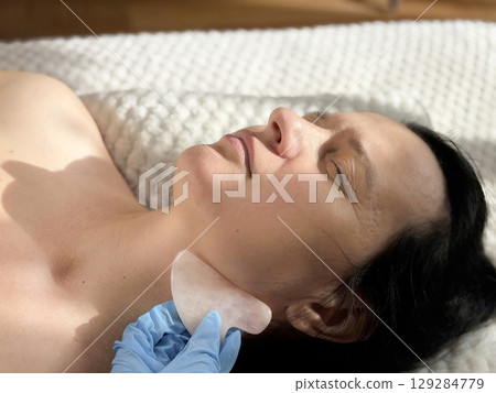 Mature woman lying on cosmetology table during professional facial massage with natural scraper. Anti aging skin care in beauty clinic. Wrinkles, skin care, beauty and wellness concept 129284779