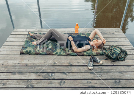 person lying flat on pier with bottle and jacket near, lake reflection and tree branches above, concept of summer rest, eco lifestyle, a young woman chooses a digital detox 129285074