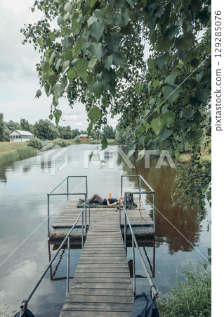 woman resting on wooden pier over calm lake with trees and rural houses in background, concept of relaxation, nature retreat, mindfulness, vertical woman resting on wooden pier over calm lake with trees and rural houses in background, concept of relaxation, nature retreat, mindfulness, vertical 129285076
