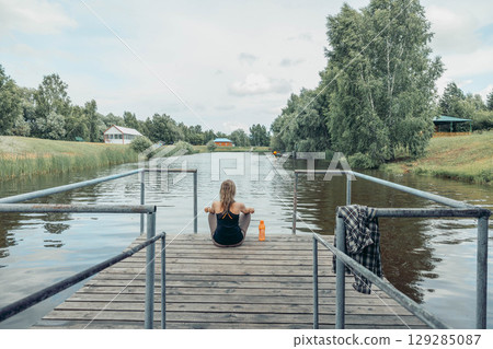 woman sitting cross-legged on wooden dock over calm river, wearing sportswear, back view, summer greenery and countryside houses, meditation, morning ritual, nature therapy woman sitting cross-legged on wooden dock over calm river, wearing sportswear, back view, summer greenery and countryside houses, meditation, morning ritual, nature therapy 129285087