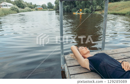 woman resting peacefully on rustic wooden dock above water, eyes closed, wearing dark clothes, rural background with green trees and boat, mindfulness, nature therapy, slow living 129285089