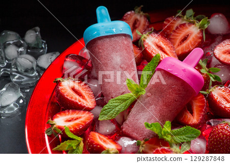 Two trawberry ice cream popsicles on red plate with ice cubes chopped strawberries and mint leaves on a black background. Strawberry ice cream popsicles. Horizontal. 129287848