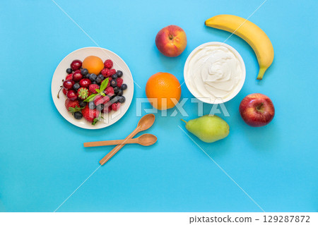 Greek yogurt around fruits orange, banana, pear, peach, apple and a plate with strawberries, raspberries, blueberries and 2 wooden spoons on light blue background. Yogurt, fruits and berries flat lay Greek yogurt around fruits orange, banana, pear, peach, apple and a plate with strawberries, raspberries, blueberries and 2 wooden spoons on light blue background. Yogurt, fruits and berries flat lay 129287872