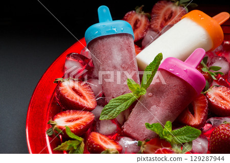 Strawberry ice cream popsicles in pink, orange, and blue hues sit atop a bed of ice, complemented by bright red strawberries and fresh mint leaves, perfect for summer enjoyment. 129287944