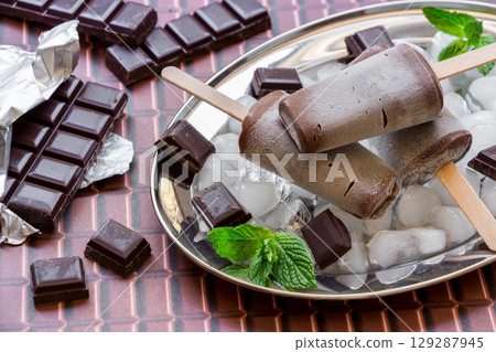 Chocolate ice cream on a stick on a silver platter with ice and pieces of chocolate mint leaves near pieces of chocolate on a chocolate background. Chocolate ice cream popsicles. Horizontal. Close. 129287945