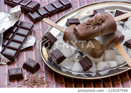 Chocolate ice cream on a stick on a silver platter with ice and pieces of chocolate near pieces of chocolate on a chocolate background. Chocolate ice cream popsicles. Horizontal. Close. Chocolate ice cream on a stick on a silver platter with ice and pieces of chocolate near pieces of chocolate on a chocolate background. Chocolate ice cream popsicles. Horizontal. Close. 129287946