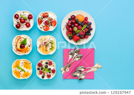 6 bowls of yogurt dressed with different ingredients flat lay near plate with berries and 6 spoons, napkins on light blue background. Six differently dressed yogurts ready to eat. Top view. 6 bowls of yogurt dressed with different ingredients flat lay near plate with berries and 6 spoons, napkins on light blue background. Six differently dressed yogurts ready to eat. Top view. 129287947