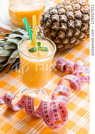 Diagonally left down a glass of pineapple smoothie with straws and mint leaf and open centimeter in the background pineapple and slice, on yellow checkered tablecloth. Close. Vertical. 129287951