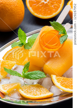 Orange sorbet ice cream decorated with a flower of orange peel on silver plate with ice cubes, orange pieces, mint near orange slices on black background. Orange sorbet ice cream popsicles. Vertical. 129287952